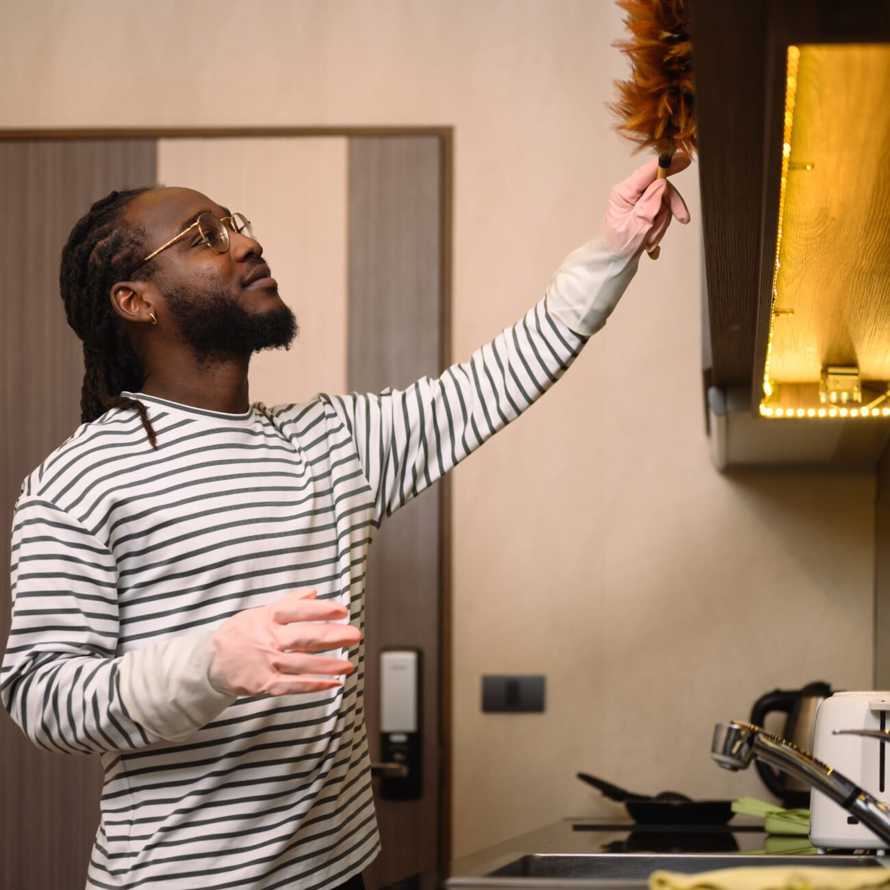 Happy bearded African man dusting shelf with a feather duster at modern kitchen.
